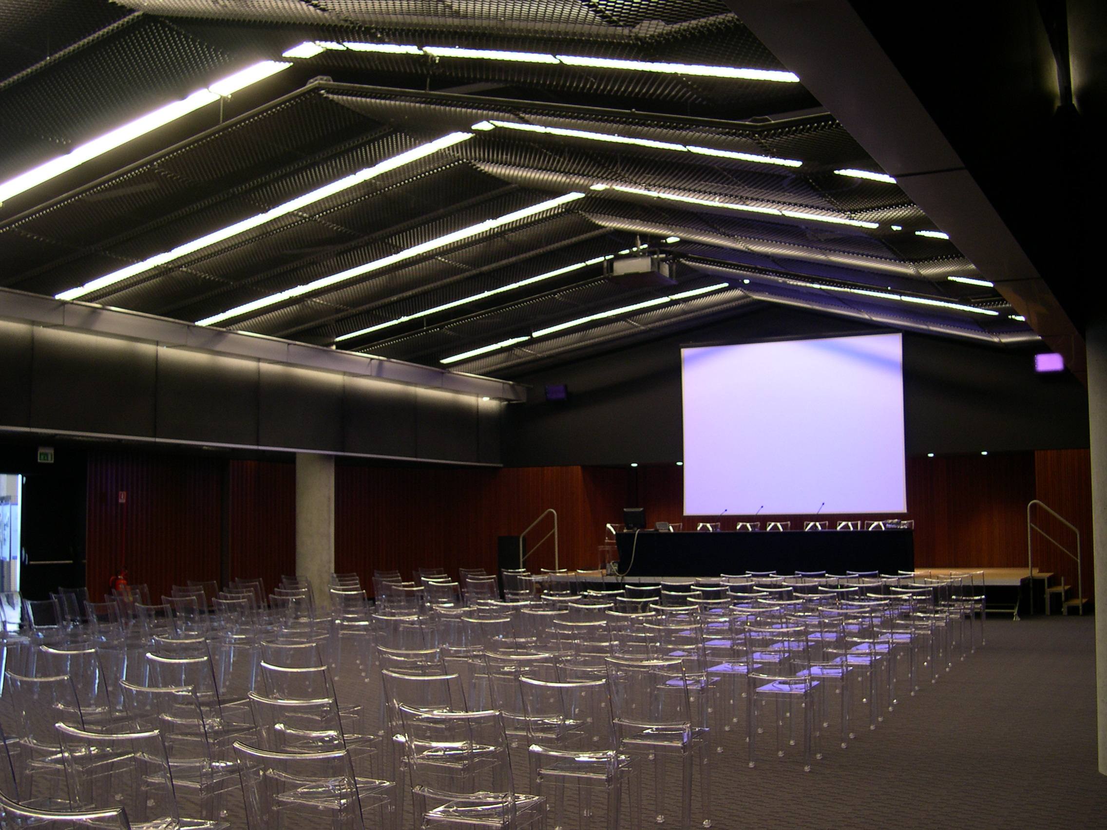 Auditorium Hypo Bank Udine con schermo di proiezione, sedie trasparenti e pannelli fonoassorbenti a parete