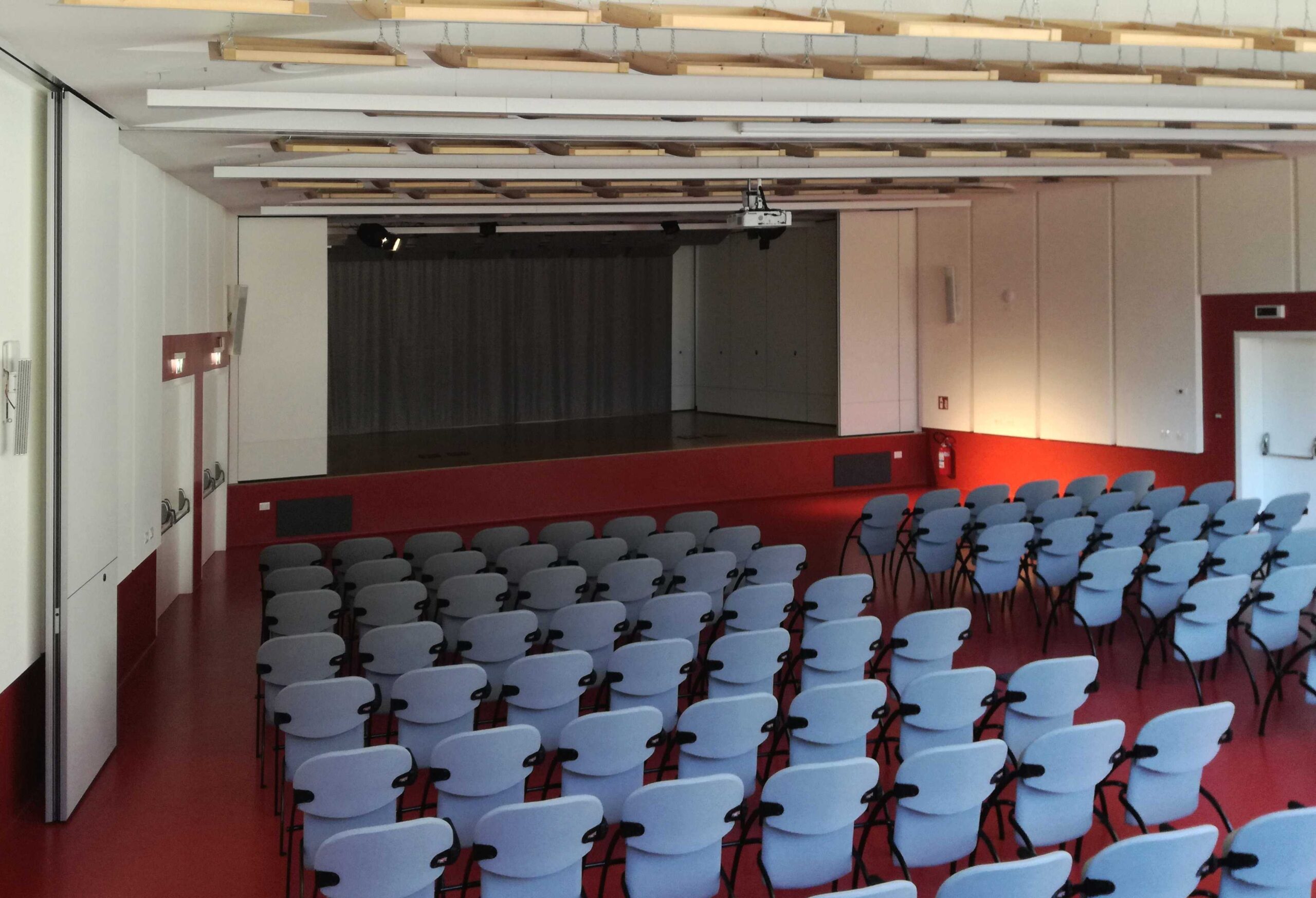 Sala polivalente del Centro Giovani di Pieve di Bono con sedie, palco e pannelli acustici a soffitto