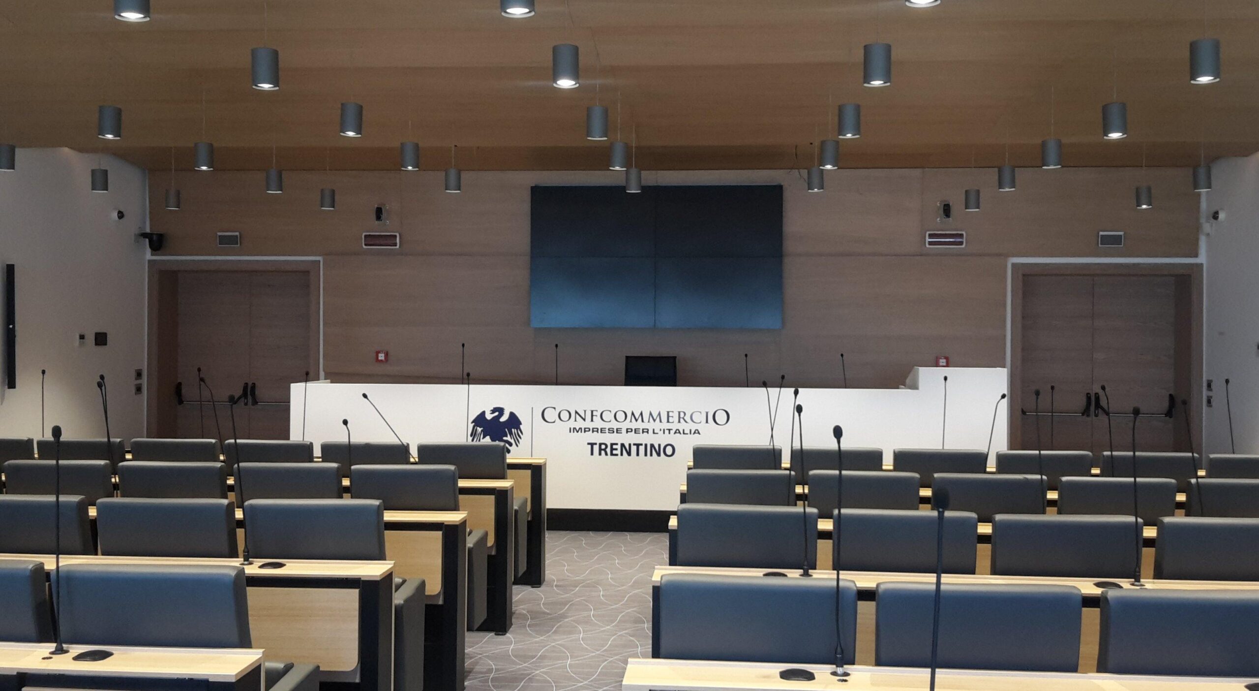 Vista frontale dell auditorium Confcommercio Trentino con videowall, microfoni e soffitto in legno