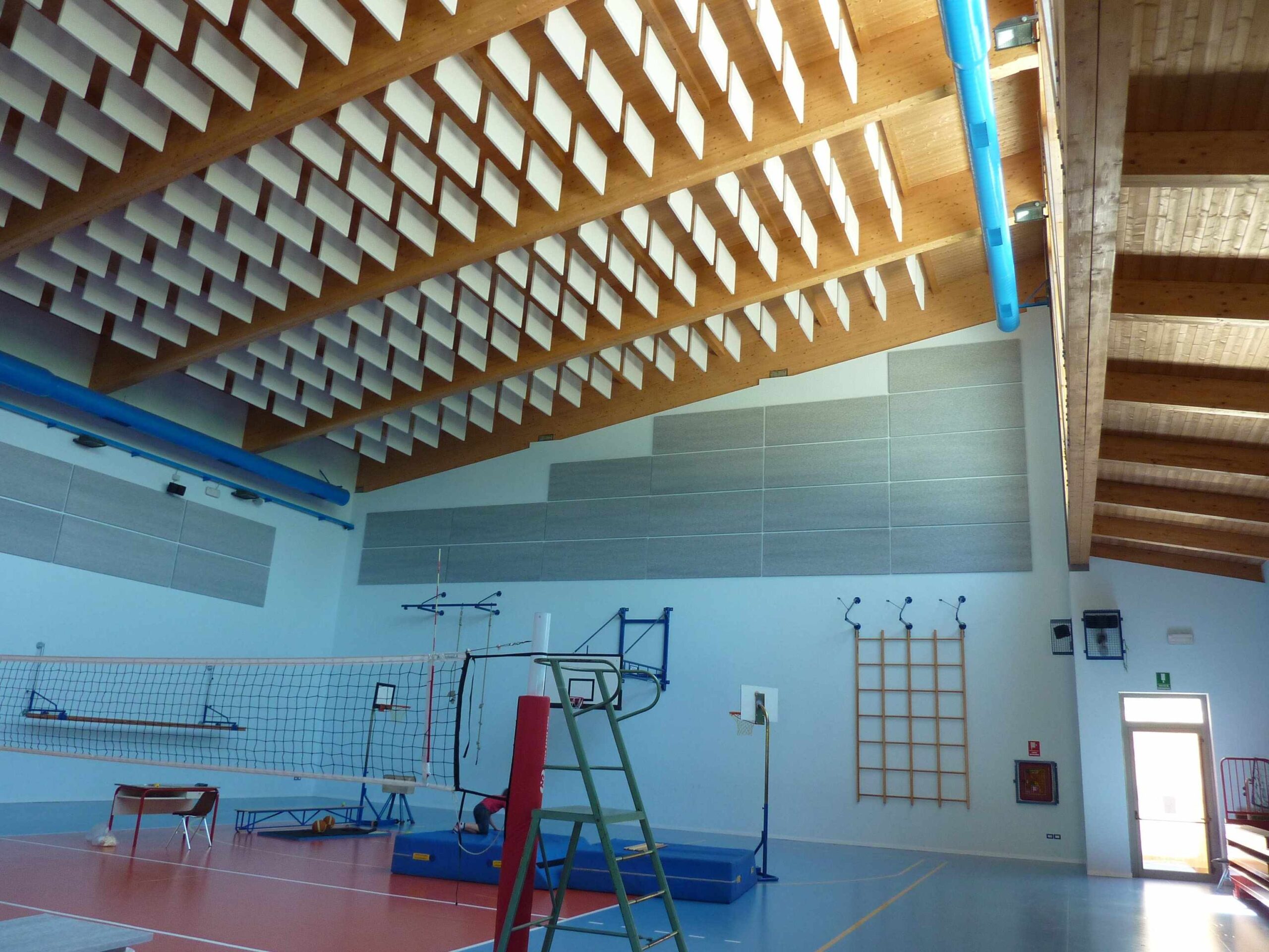 Interno di palestra con pannelli acustici a soffitto, rete da pallavolo e attrezzature sportive