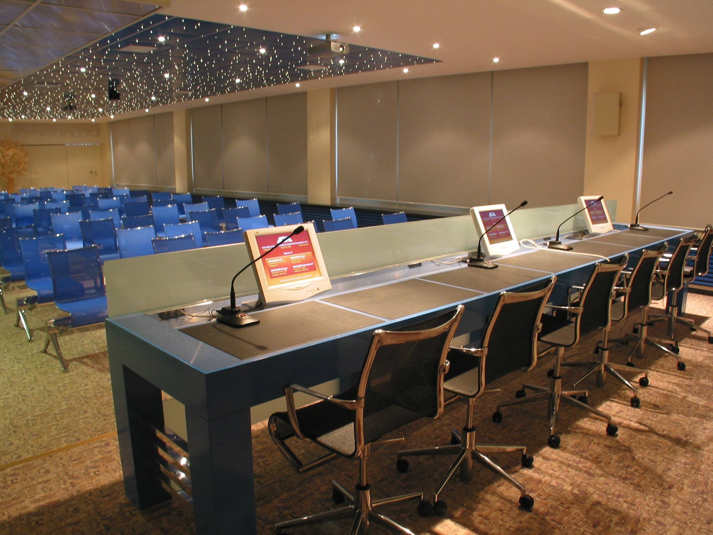 Tavolo relatori con microfoni e monitor nella sala meeting Foppa Pedretti con sedute blu