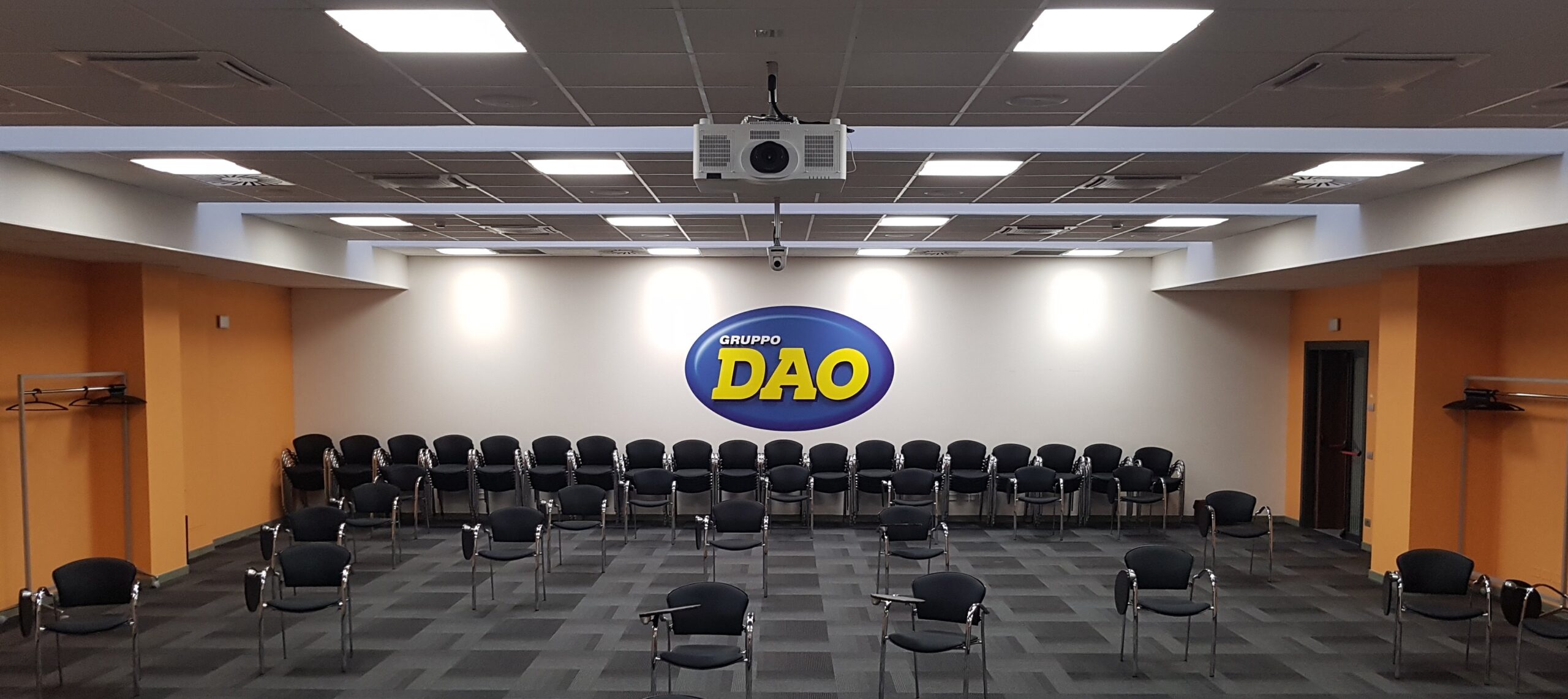 Sala assemblee Gruppo DAO con logo a parete, proiettore a soffitto e sedie nere disposte in file