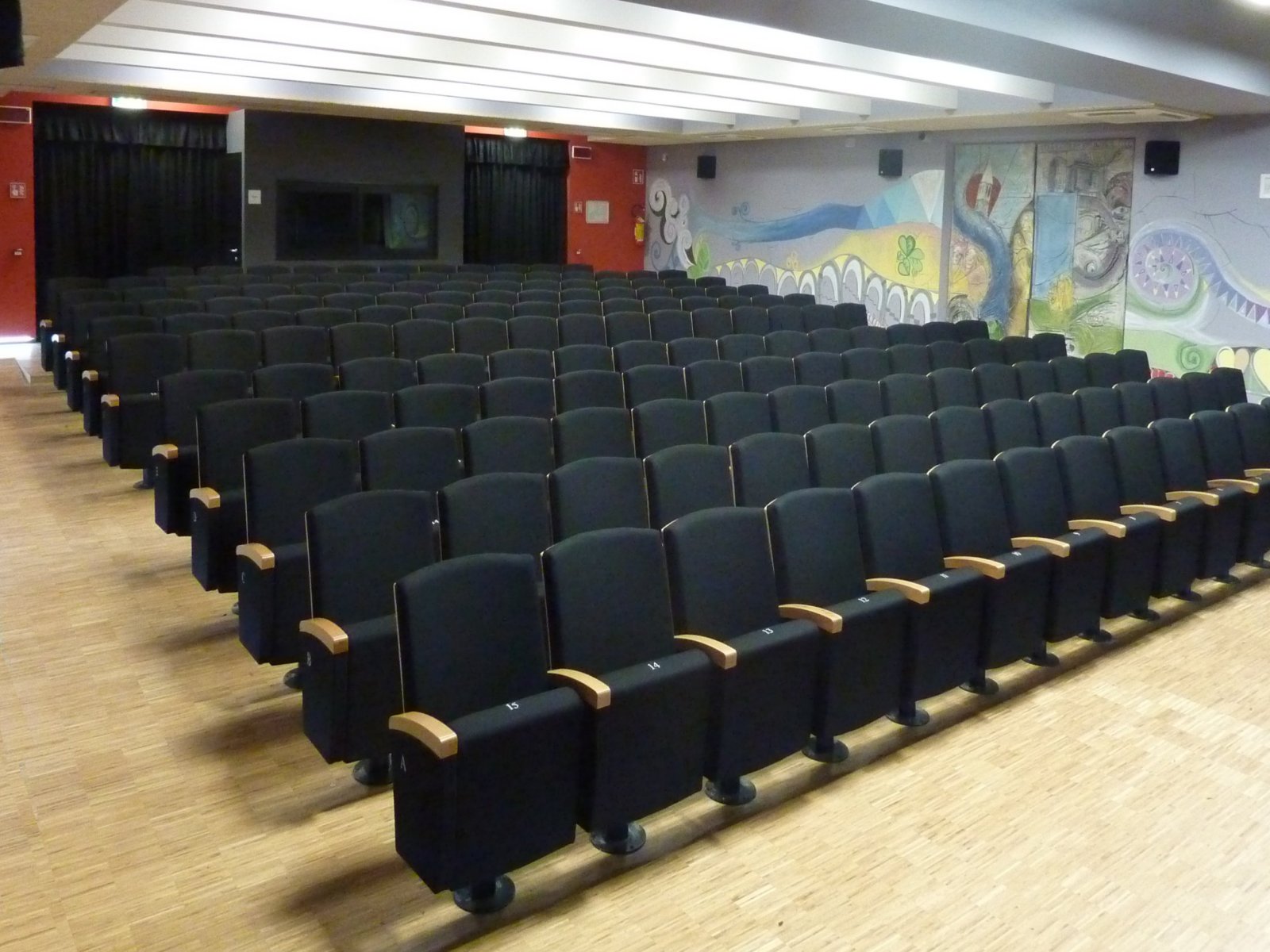 Poltrone imbottite con braccioli in legno e murales colorato nella sala polifunzionale di Gardolo