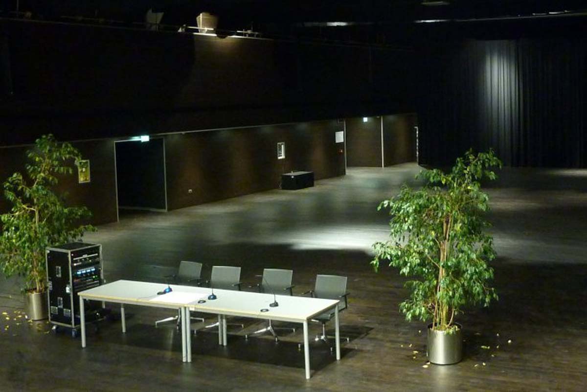 Palcoscenico della sala Sanbapolis con tavolo conferenze, microfoni e piante decorative