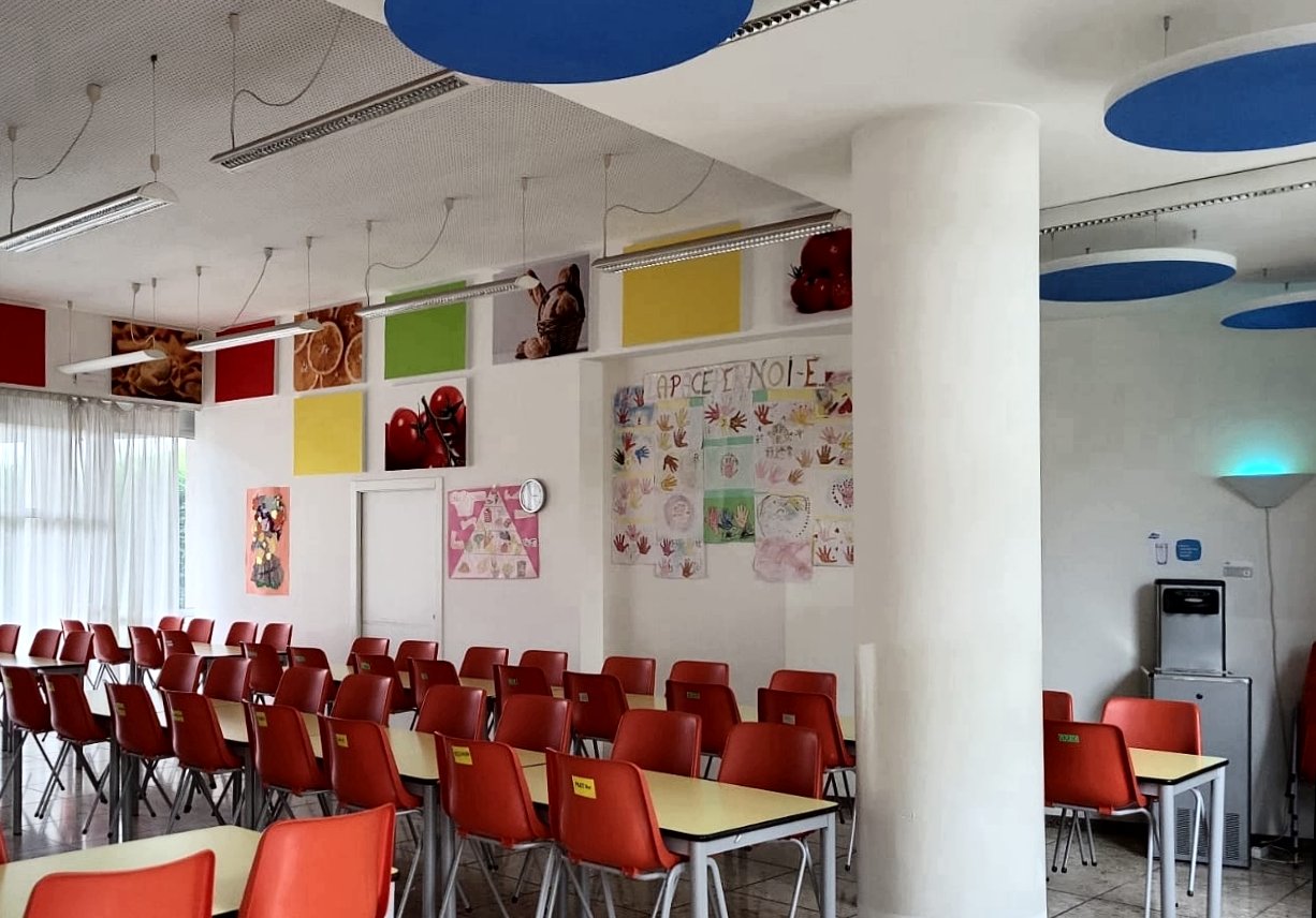 Mensa scolastica con sedie rosse, pannelli acustici colorati e dischi blu a soffitto a Ville d'Anaunia