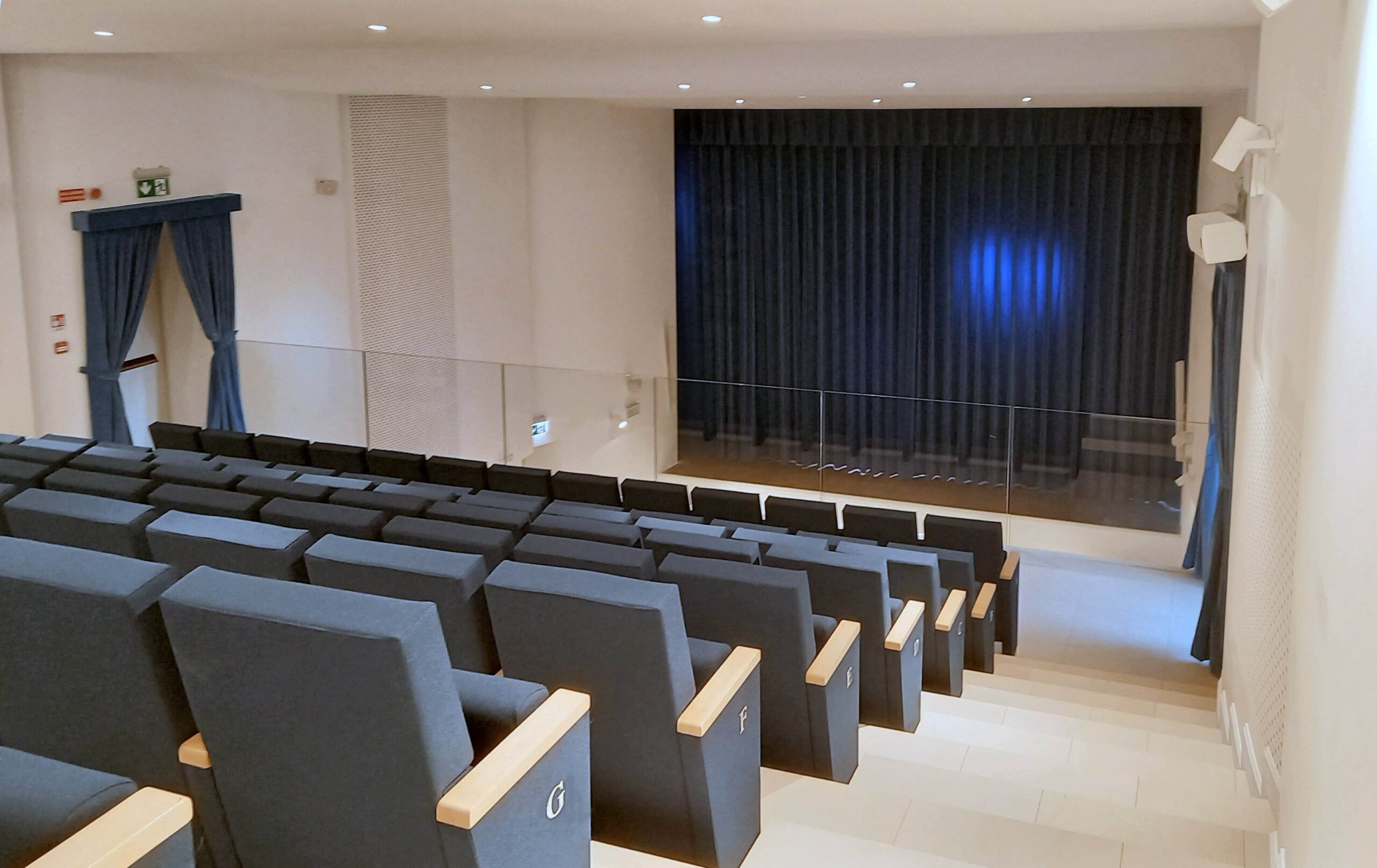 Platea del teatro di Calcinato con poltrone grigio-blu, palcoscenico con sipario scuro e diffusori audio